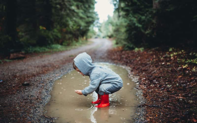 A young boy in a hoodie and bright red rubber boots squats in a muddy puddle