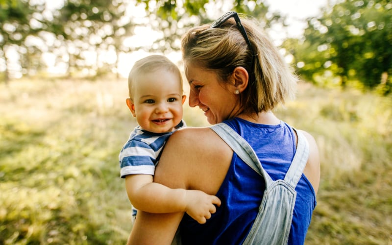 A mother carries her smiling toddler in a meadow